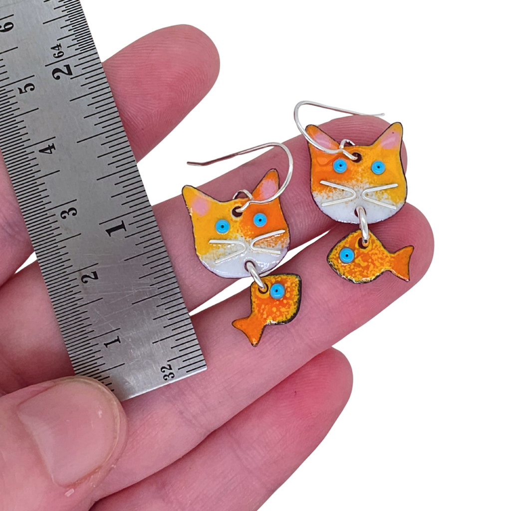 Cat-shaped earrings with fish details held by a hand next to a ruler for size reference.