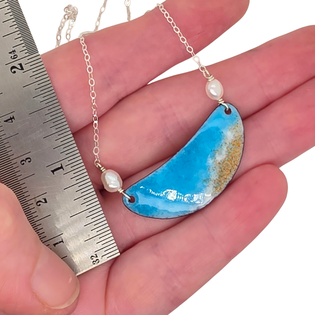 Jewelry with a blue pendant and pearl beads held by a hand next to a ruler.