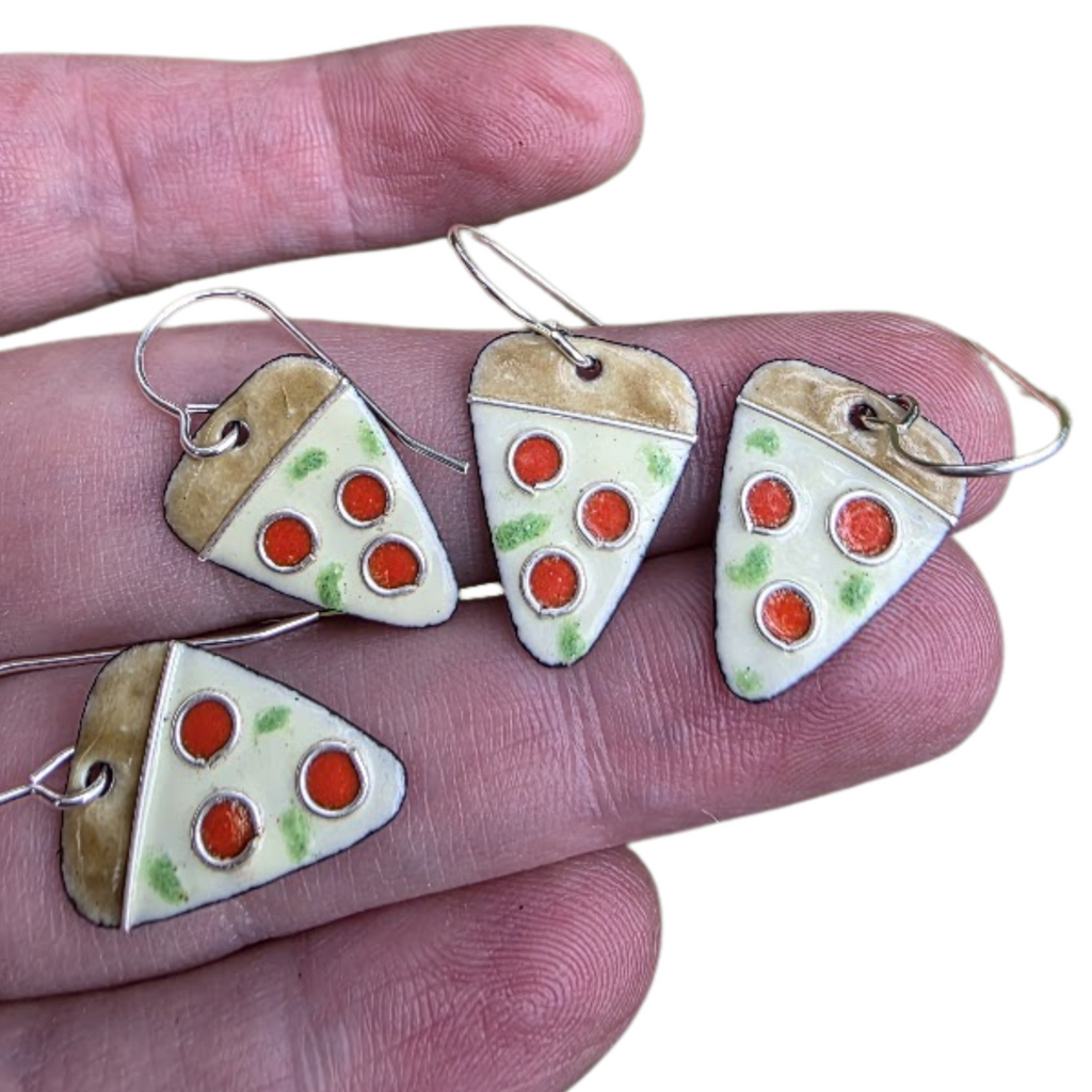 Pizza slice earrings held between fingers on a white background.  Earrings are artisan made from glass enamel fused to copper with fine silver and sterling silver accents.