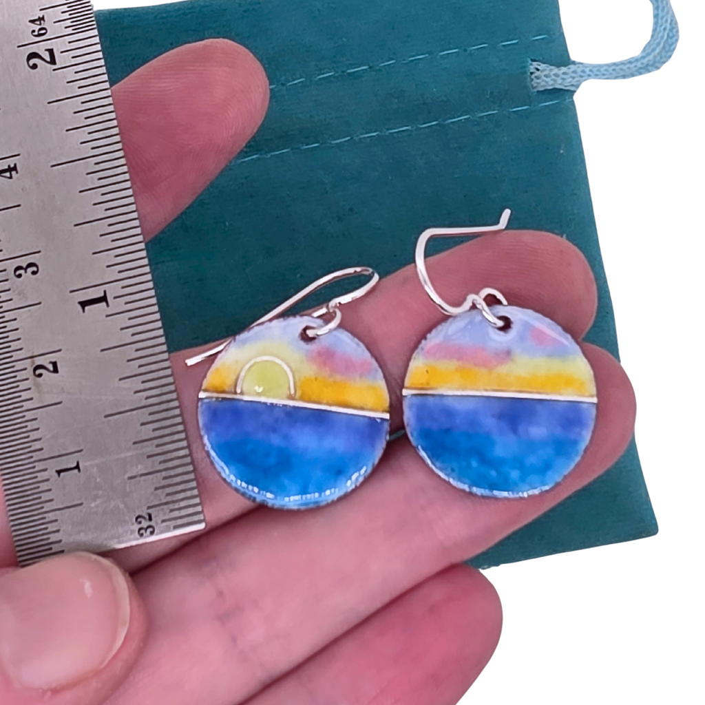 Colorful glass enamel earrings held between fingers with a ruler for scale against a teal background