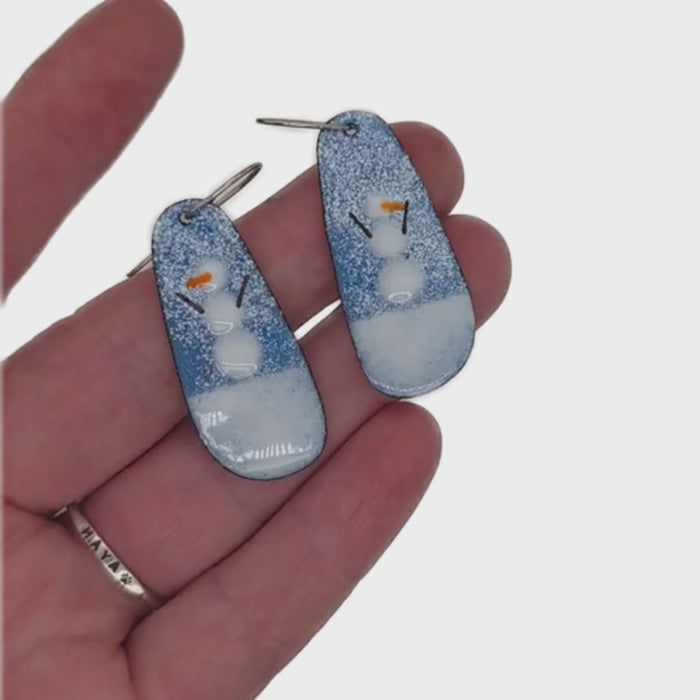 A pair of handmade teardrop-shaped drop earrings featuring a snowman design. The earrings have a textured blue upper section with white speckles representing snow, a white lower section representing snow, and a small snowman with orange carrot nose and  black stick arms.